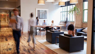 A group of people sitting at a table in an office, some engrossed in conversation while others are focused on their laptops.