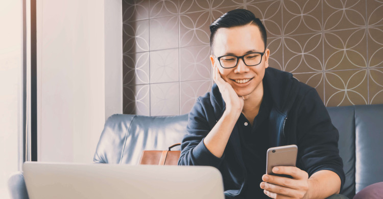 front view of man in causal clothes sitting on sofa smiling and holding mobile phone with laptop in view