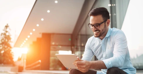 man smiling with glasses wearing smart clothes sitting outside on steps with tablet in hand