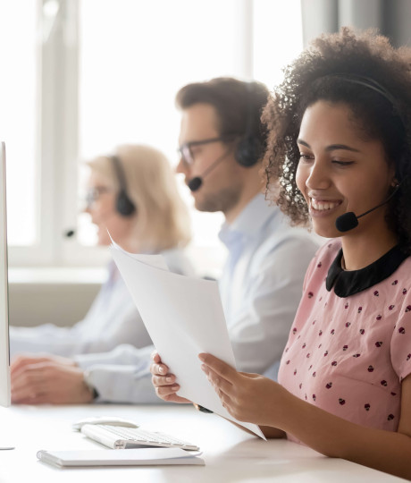 woman at a desk in a call center with headset on holding documents with two people in background