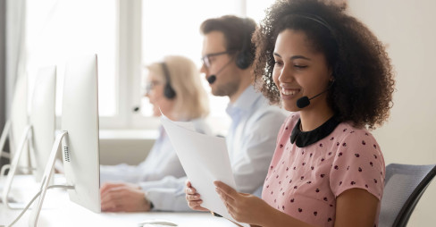 woman at a desk in a call center with headset on holding documents with two people in background