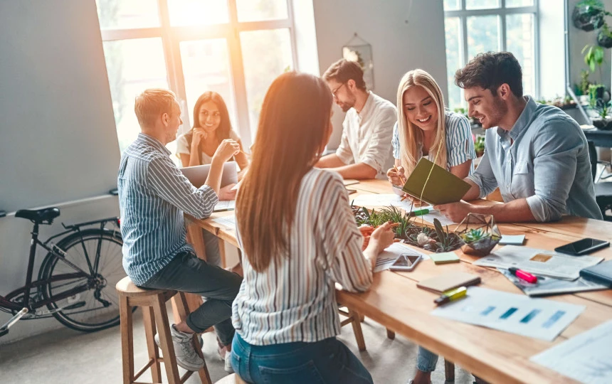 six people sitting in a communal area smiling and interacting with office material on the table