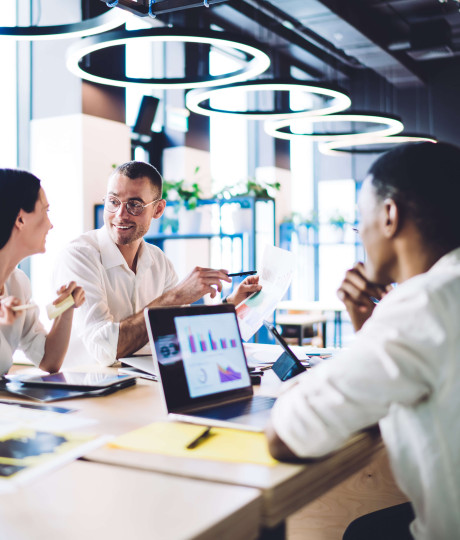four people in smart clothing sitting in a office communal area with laptops and documents