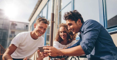 three people sitting outside smiling and looking at a mans mobile phone
