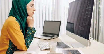 woman in hijab analysing data on apple mac with laptop and notes on the desk