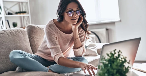 woman with glasses wearing casual clothes sitting with legs crossed on sofa looking at a laptop