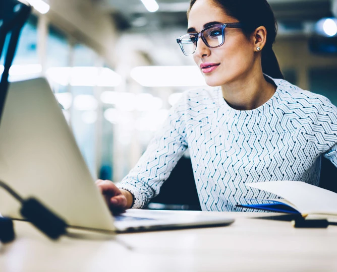 woman with glasses looking at laptop with pen in hand