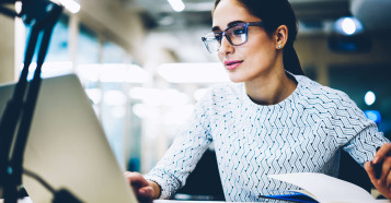 woman with glasses looking at laptop with pen in hand