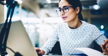 woman with glasses looking at laptop with pen in hand
